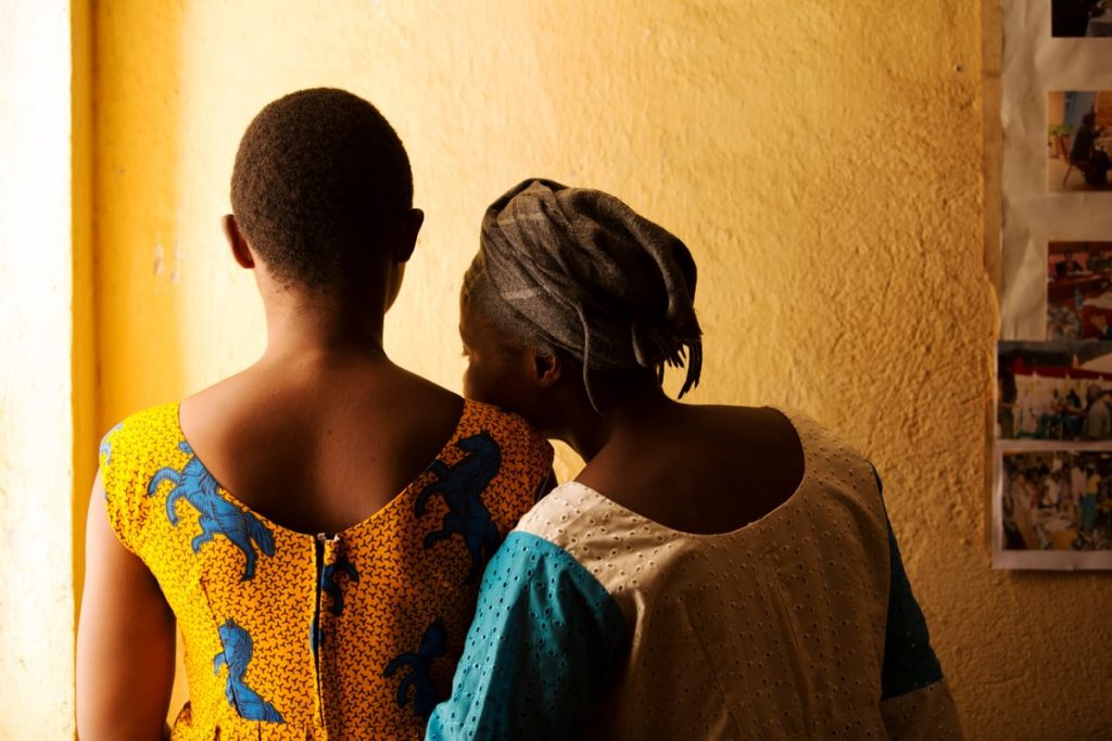 Two women stood with their backs to the camera, one with her head on the others shoulder.