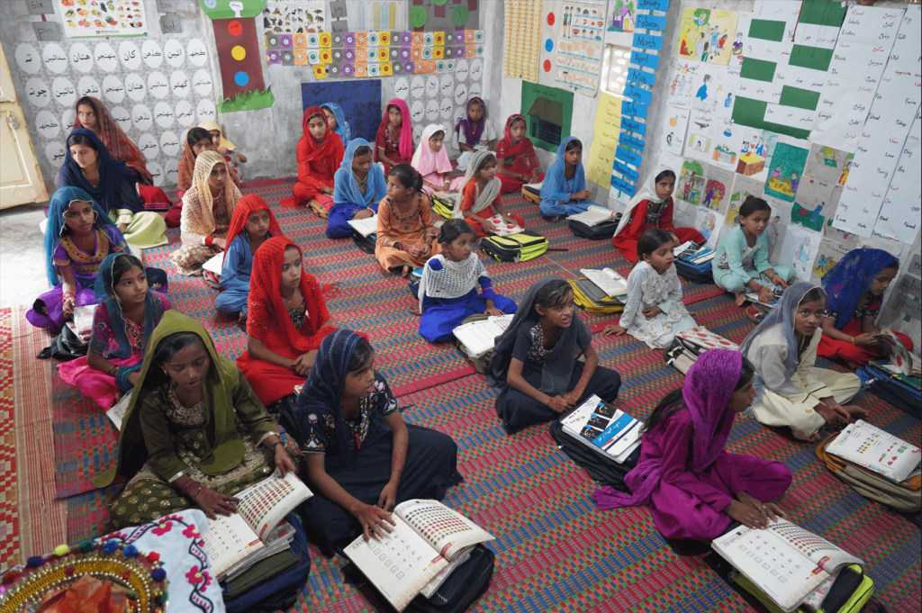 Girls in colourful clothing sat on a rug on the floor with books open, and learning materials all over the walls. 